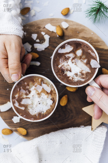 Woman and man holding a cup of vegan almond milk hot chocolate topped with coconut butter, coconut chips and almonds.
