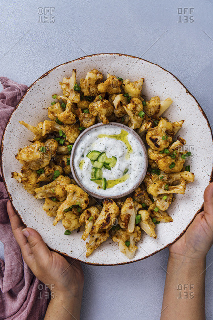 Hands holding roasted spicy cauliflower wings in a ceramic bowl served with a small bowl of yogurt cucumber dip