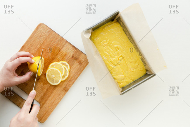 Hands cutting lemons on a wooden cutting board next to a loaf pan of yellow batter