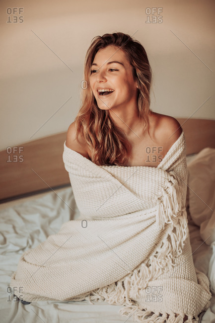 Young smiling woman wrapped in blanket on bed with her bare shoulders exposed