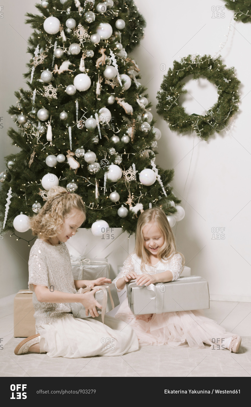 Two little girls sitting in front of Christmas tree opening Christmas presents