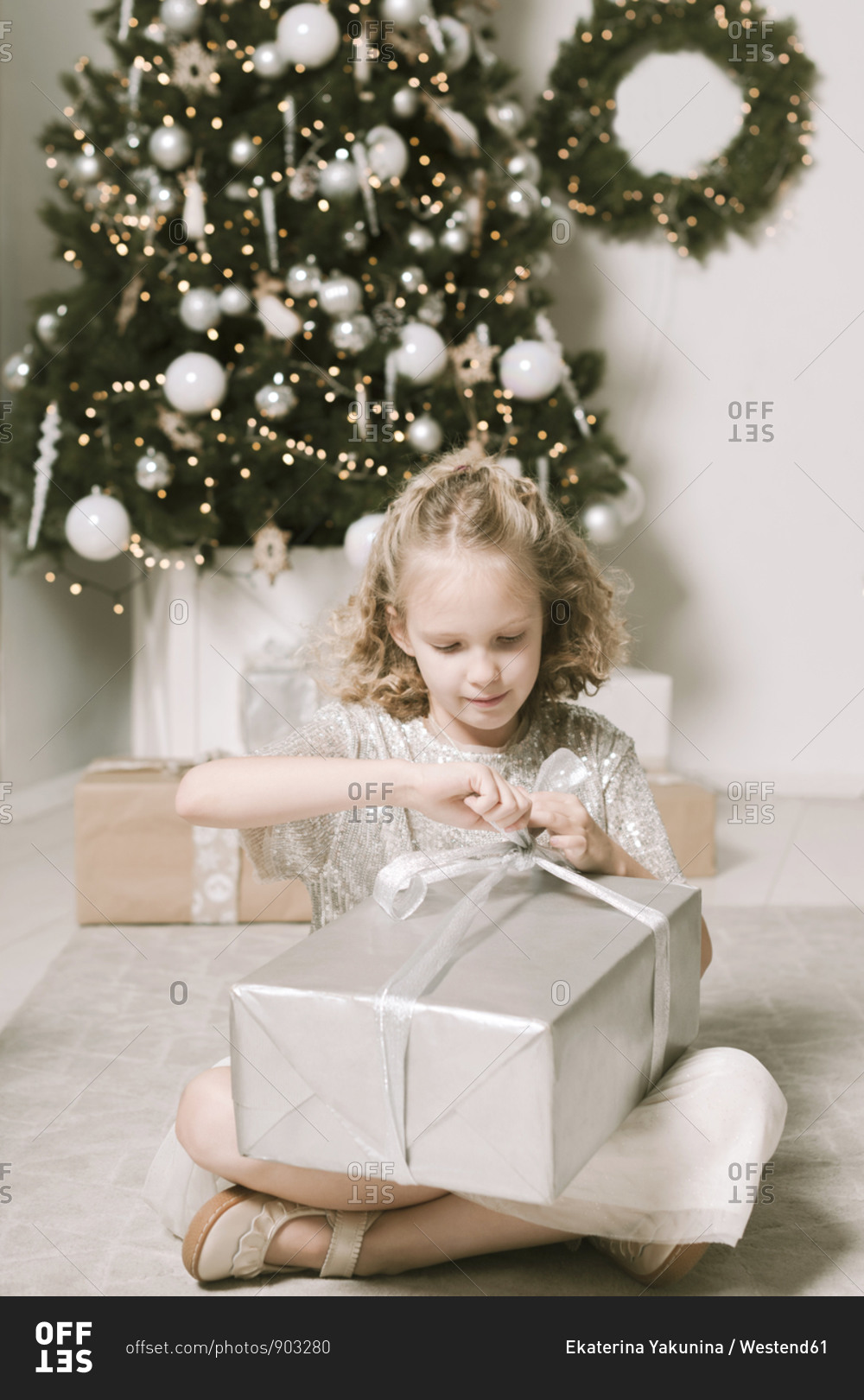 Portrait of blond little girl sitting in front of Christmas tree opening Christmas present