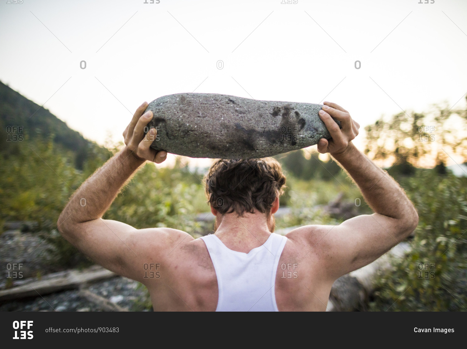 Rear view of man lifting rock above head during an outdoor workout.