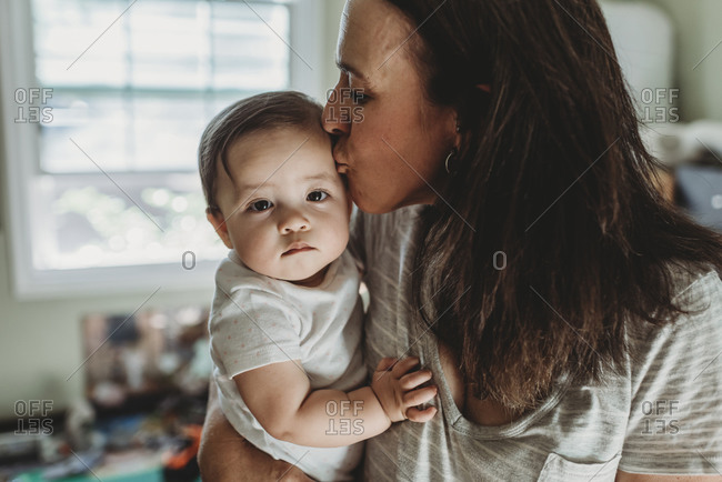 Mid-40s mom kissing baby daughter with big brown eyes in front of window