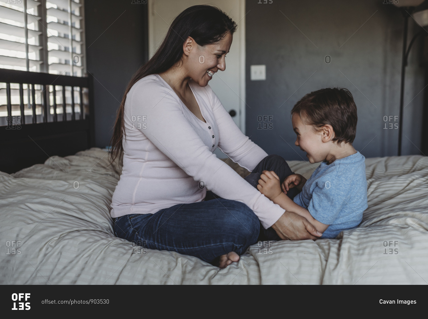 Laughing mid-30s mom tickling 5 yr old son on bed near window