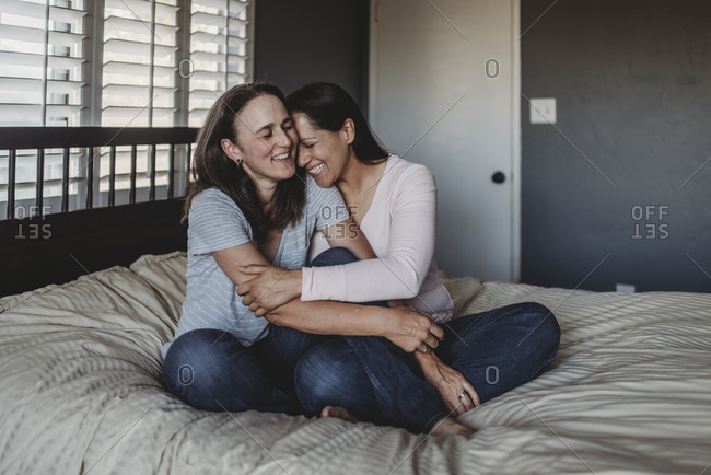 Laughing same sex couple hugging while sittings on bed near window