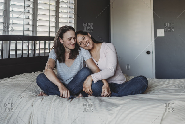 Smiling same sex couple snuggling on bed near window with shutters