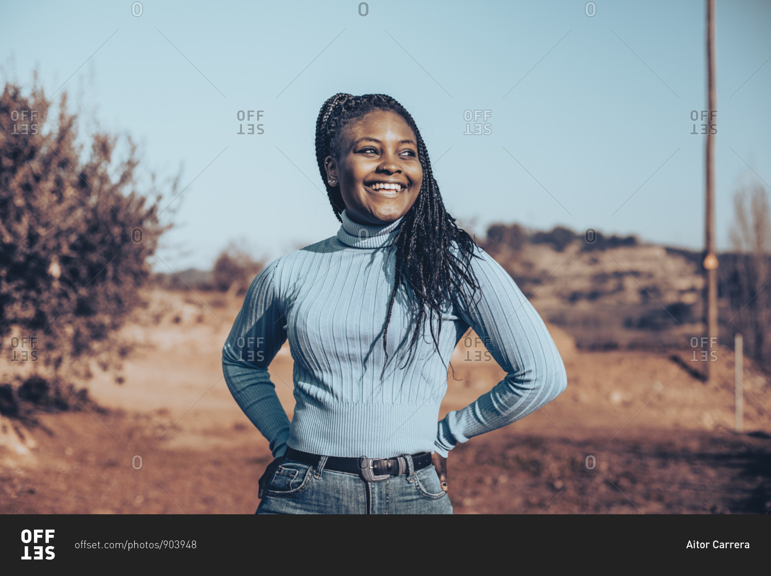 Smiling woman with braids wearing a blue turtleneck sweater standing in a rural setting