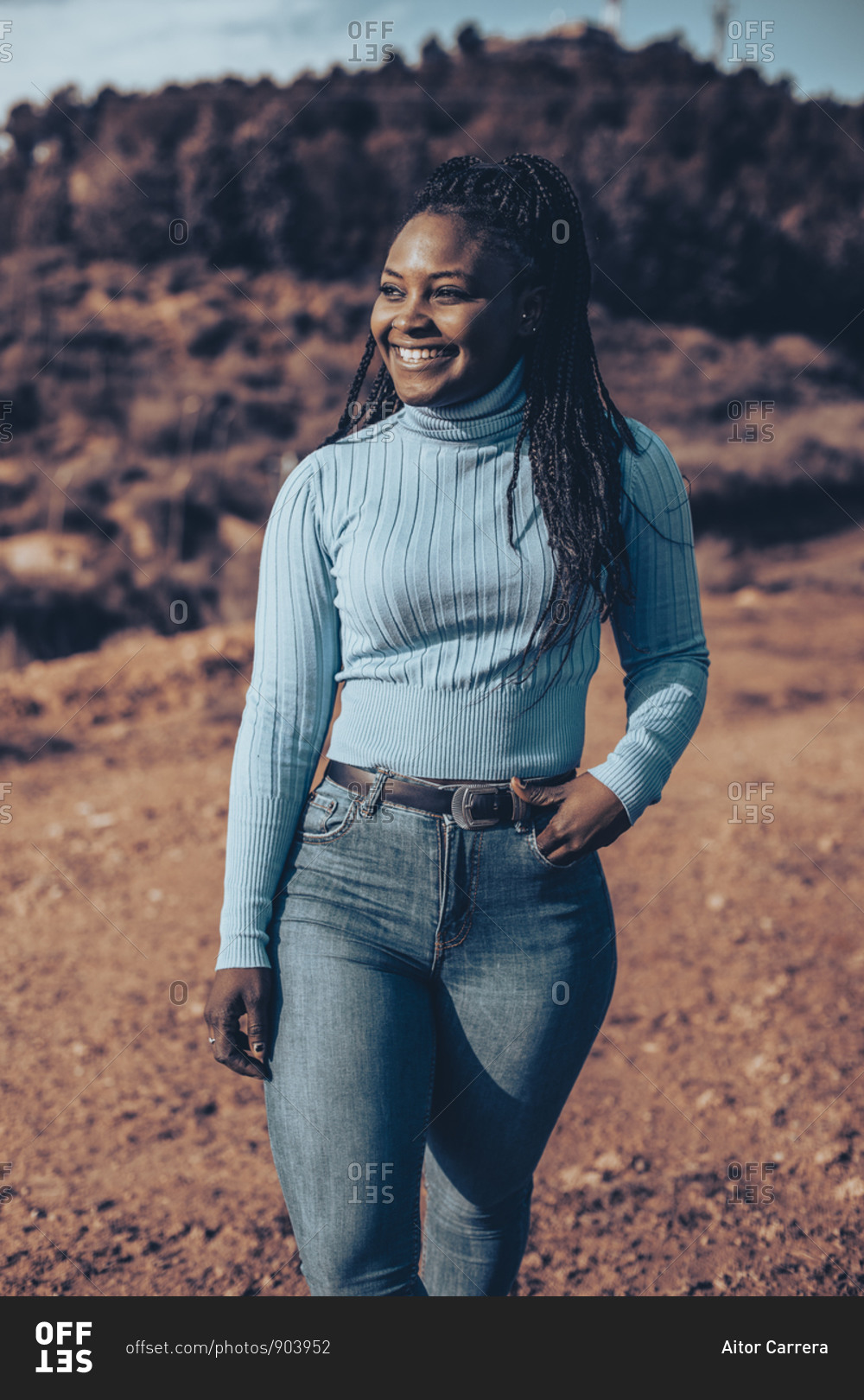 Side view of a woman with braids wearing a blue turtleneck sweater standing in a rural setting