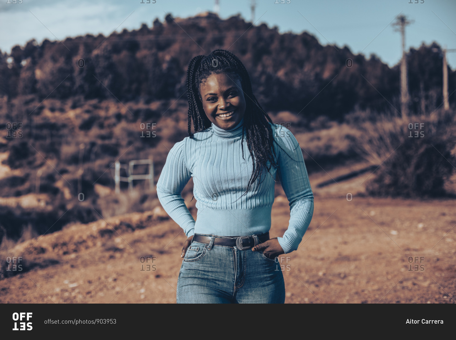 Smiling woman with braids wearing a blue turtleneck sweater\
standing in a rural setting stock photo - OFFSET