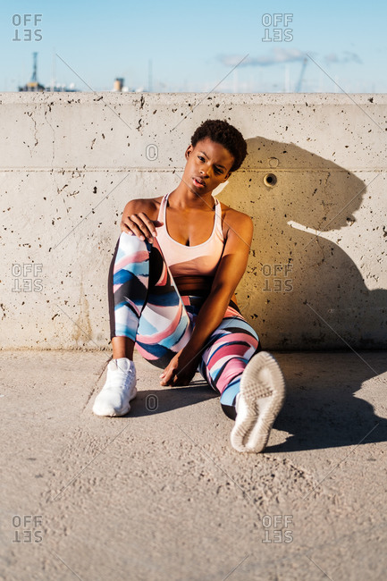 African American female athlete in flowered sport clothes and white sneakers looking at camera while sitting alone on concrete wall in sunbeams and resting after training in city