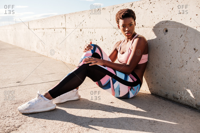 African American female athlete in flowered sport clothes and white sneakers looking at camera while sitting alone on concrete wall in sunbeams and resting after training in city