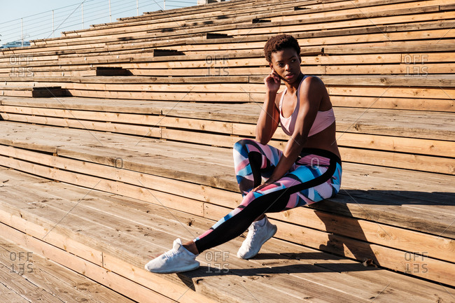 Slim African American young woman in colorful sport clothes and white sneakers looking away with curiosity and contemplating while sitting alone on brown wooden stairs on stadium