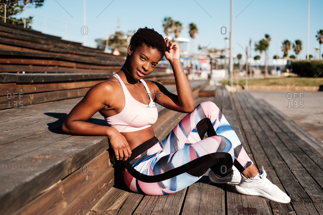 Slim African American young woman in colorful sport clothes and white sneakers looking at camera with curiosity and contemplating while sitting alone on brown wooden stairs on stadium