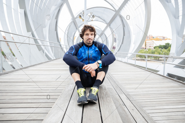 Male jogger in workout clothes sitting on wooden ground of enclosed bridge resting after workout looking at camera