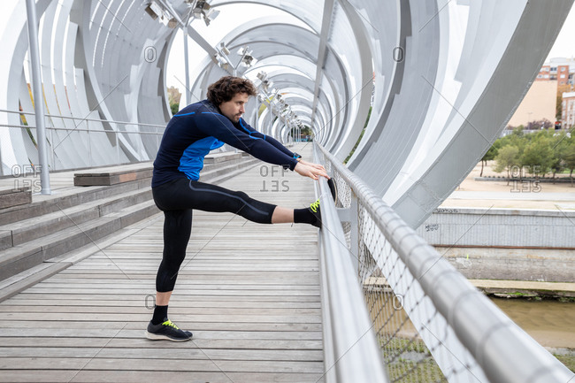 Side view of mature jogger standing on boardwalk and performing basic stretching exercises on bar at river bank at daytime