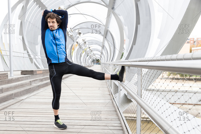 Side view of mature jogger standing on boardwalk and performing basic stretching exercises on bar at river bank at daytime