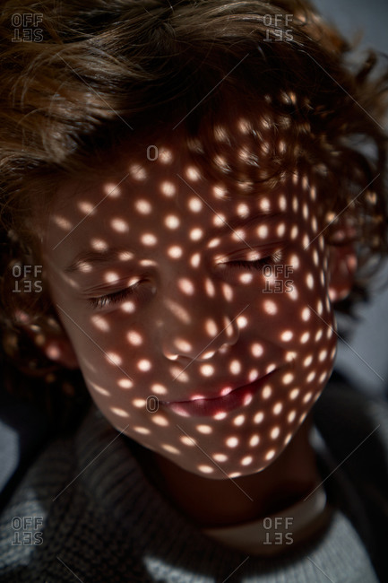 Kid slightly smiling while lying on back under source of light cast through perforated object and almost napping