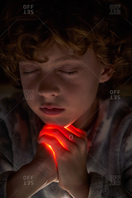 Cute child with closed eyes clasping hands with bright light and praying at night with