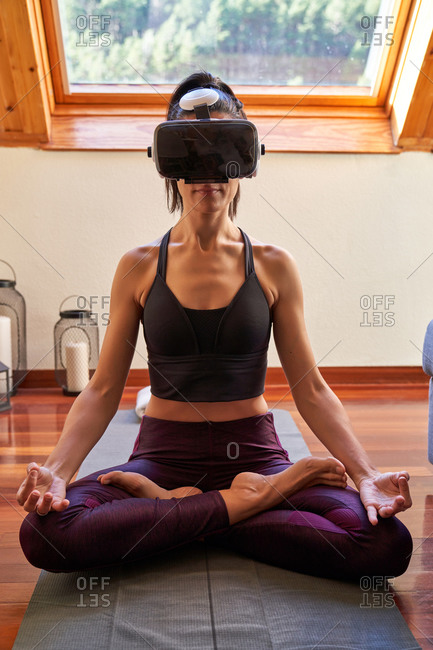Adult female in VR glasses sitting on mat in Lotus pose and meditating while doing yoga and exploring virtual reality in morning at home