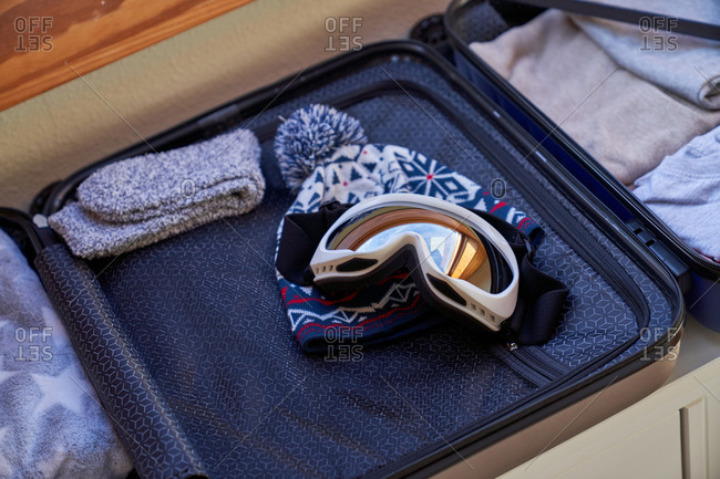 Ski goggles and hat placed in open luggage on top of drawer under window during winter vacation preparation at home