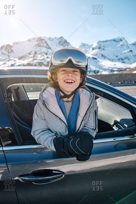 Delighted cute boy in helmet with goggles smiling looking at camera while peeking out vehicle window on sunny day on ski resort