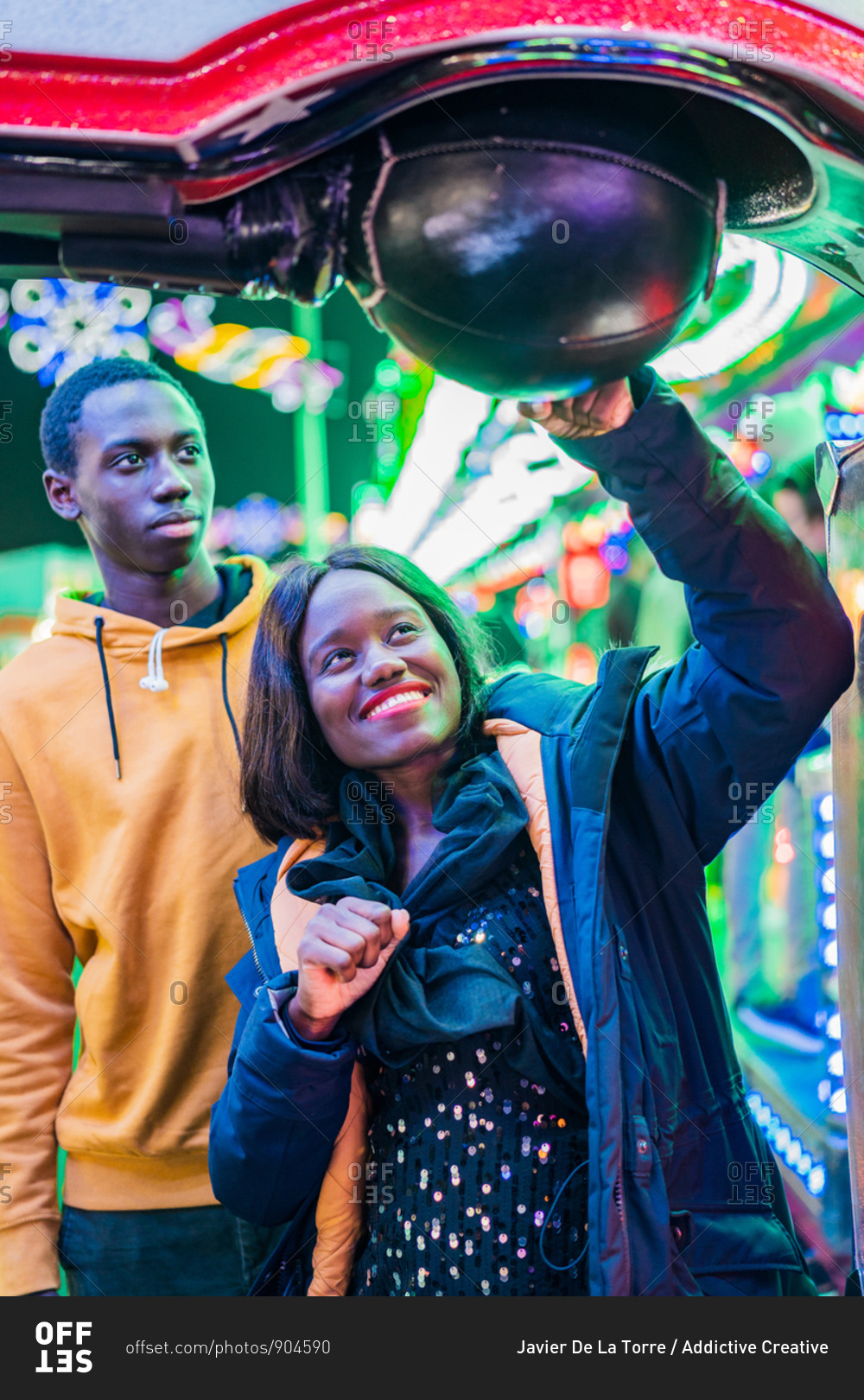Happy black woman smiling and punching ball while playing arcade game with boyfriend at night on funfair