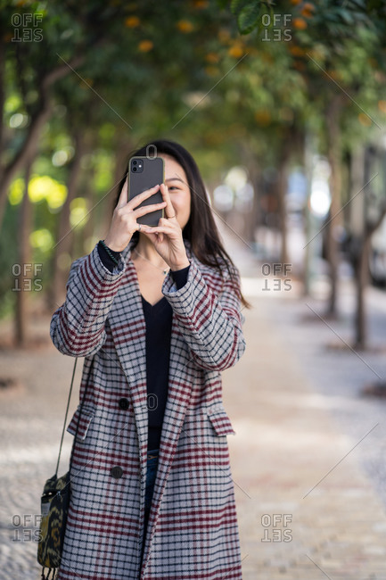 Asian female in checkered jacket with handbag focusing on screen and taking shot with smartphone while standing against blurred alley with rows of orange trees in Spain