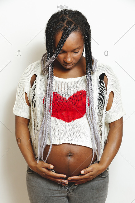 Cheerful young pregnant black female with braids wearing stylish white blouse with red heart touching belly and looking away with smile while standing against white wall