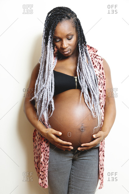 Cheerful young pregnant black female with braids wearing stylish white blouse with red heart touching belly and looking away with smile while standing against white wall