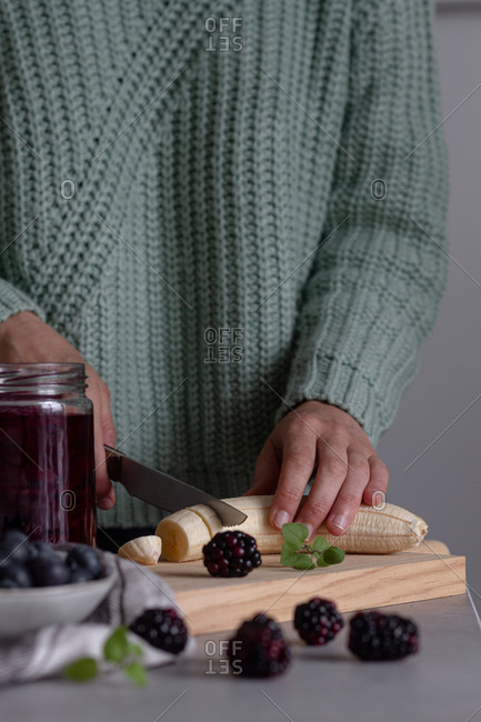 Crop person cutting with knife fresh banana on wooden cutting board while preparing healthy vitamin smoothie with blackberry and blueberry at home