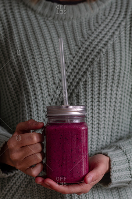 Crop person in casual knitted sweater holding transparent jar with metal lid and plastic straw of tasty vitamin pink beverage of sweet fresh fruits and berries while having healthy appetite breakfast