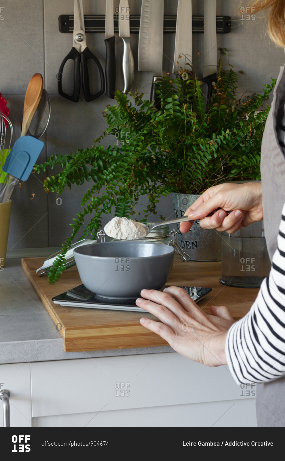Crop hands of faceless woman in apron adding spoon of flour into bowl on scale while cooking in modern kitchen