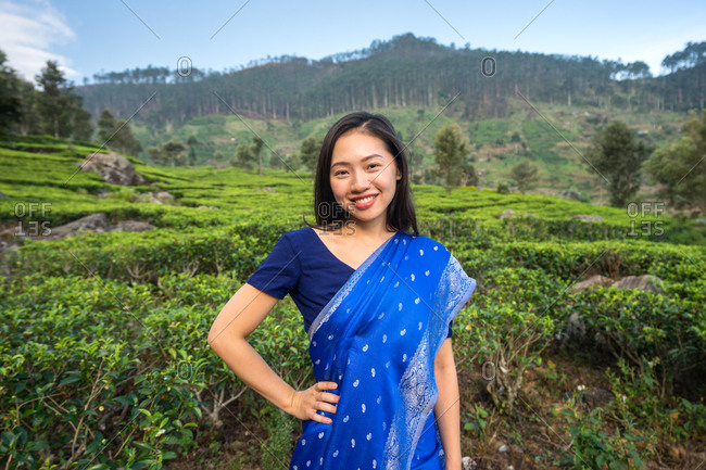 Cheerful youthful attractive ethnic female in blue traditional clothes looking at camera while standing on tea meadows in Haputale in Sri Lanka