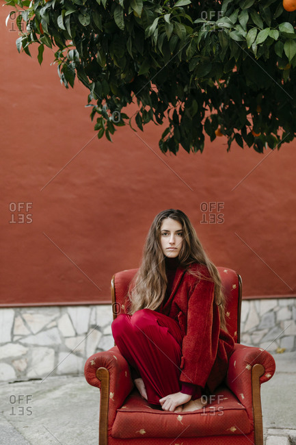 Portrait of young woman with long brown hair dresses in red crouching on red lounge chair
