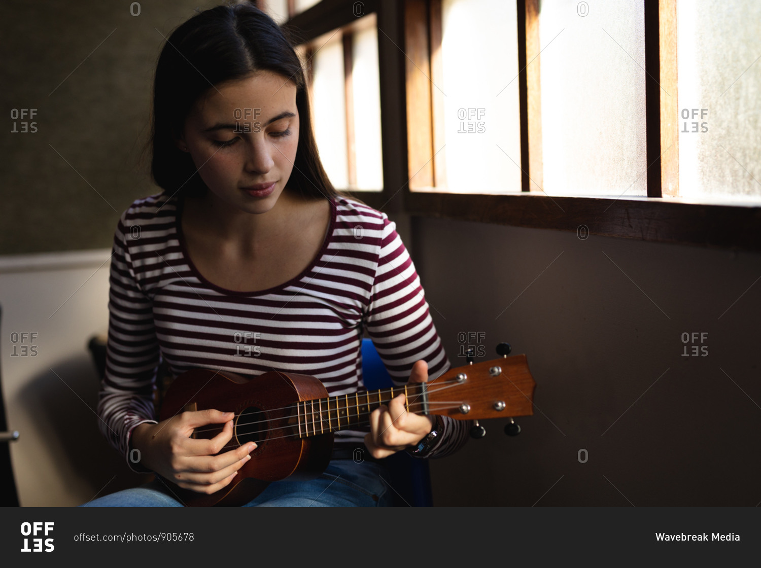 Portrait of a Caucasian musician teenage girl sitting by a window, looking down and playing a ukulele in a high school
