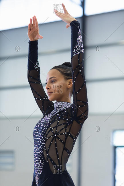 Side view close up of teenage mixed race female gymnast performing at the gym, standing with arms up, wearing black leotard