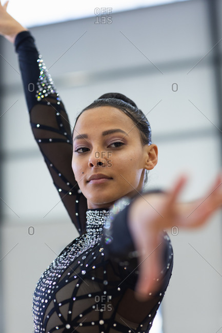 Front view close up of teenage mixed race female gymnast performing at sports hall, standing with arm up, wearing black leotard