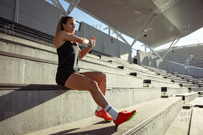 Side view of a Caucasian female athlete practicing at a sports stadium, stretching at a stand. Track and Field Sports Training in Stadium.