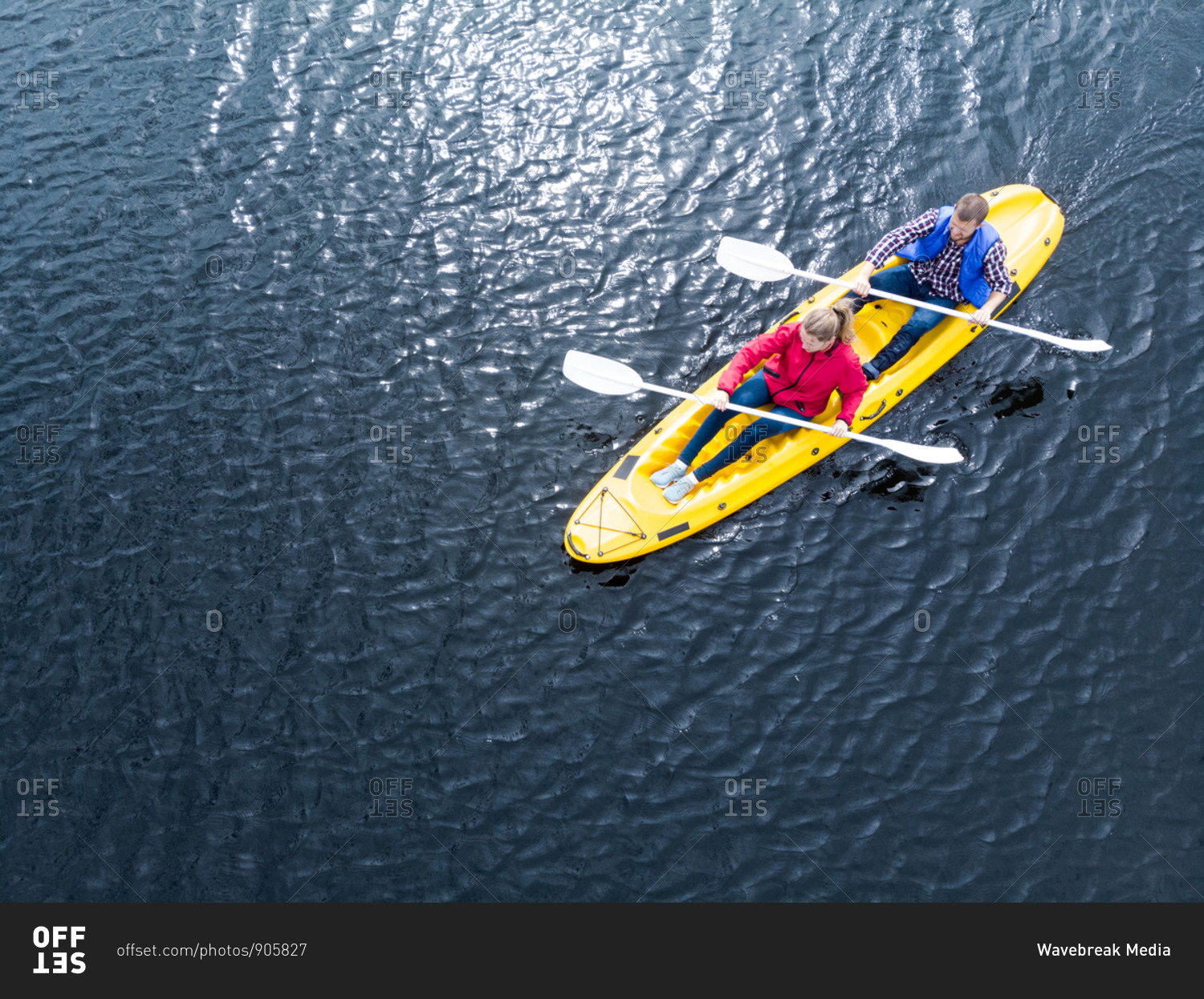 Drone shot of a Caucasian couple kayaking together in a\
yellow boat on calm water, both paddling with double-ended oars\
stock photo - OFFSET