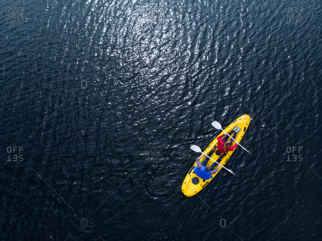 Drone shot of a Caucasian couple kayaking together in a yellow boat on calm water, both paddling with double-ended oars