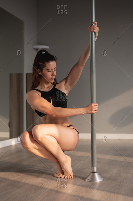 Front view of a fit attractive Caucasian woman enjoying pole dance training at a studio, holding the pole with both hands, crouching