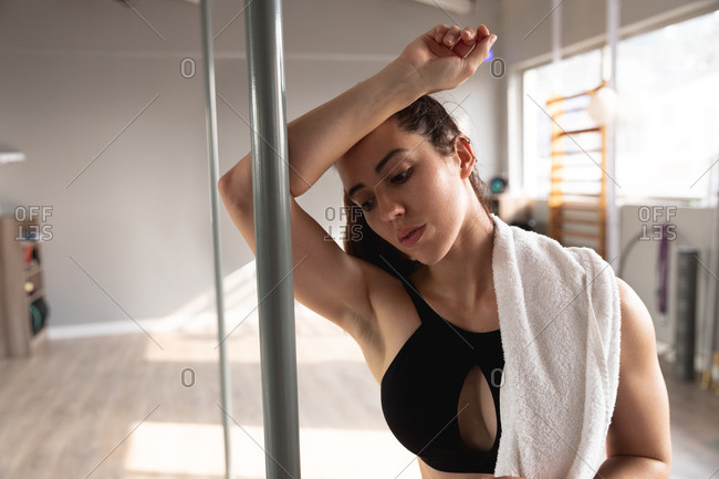 Front view of a fit attractive Caucasian woman enjoying pole dance training at a studio, taking a break, leaning on the pole with a towel on her shoulder