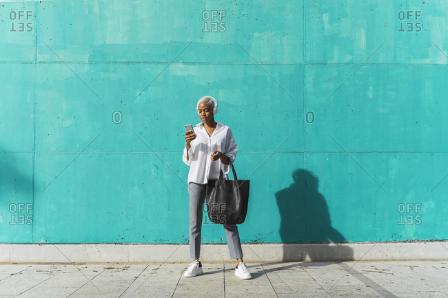 Businesswoman with headphones- standing in front of teal wall- using smartphone