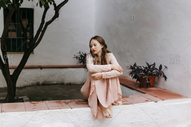 Portrait of melancholic young woman sitting barefoot on a wall hugging her knees