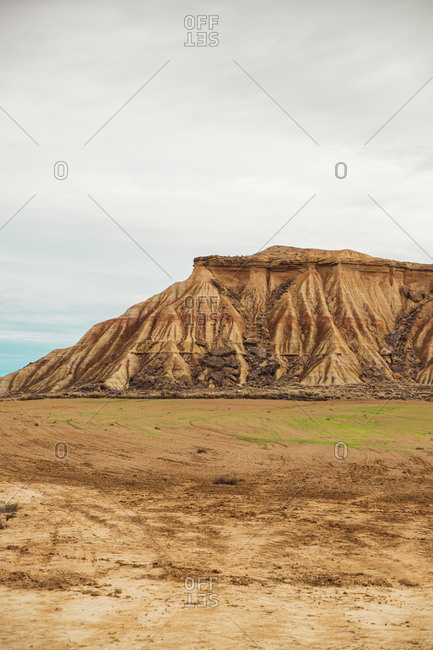 Empty field with green sparse vegetation and brown powerful cliff on background under cloudy blue sky in Bardenas Reales, Navarre, Spain