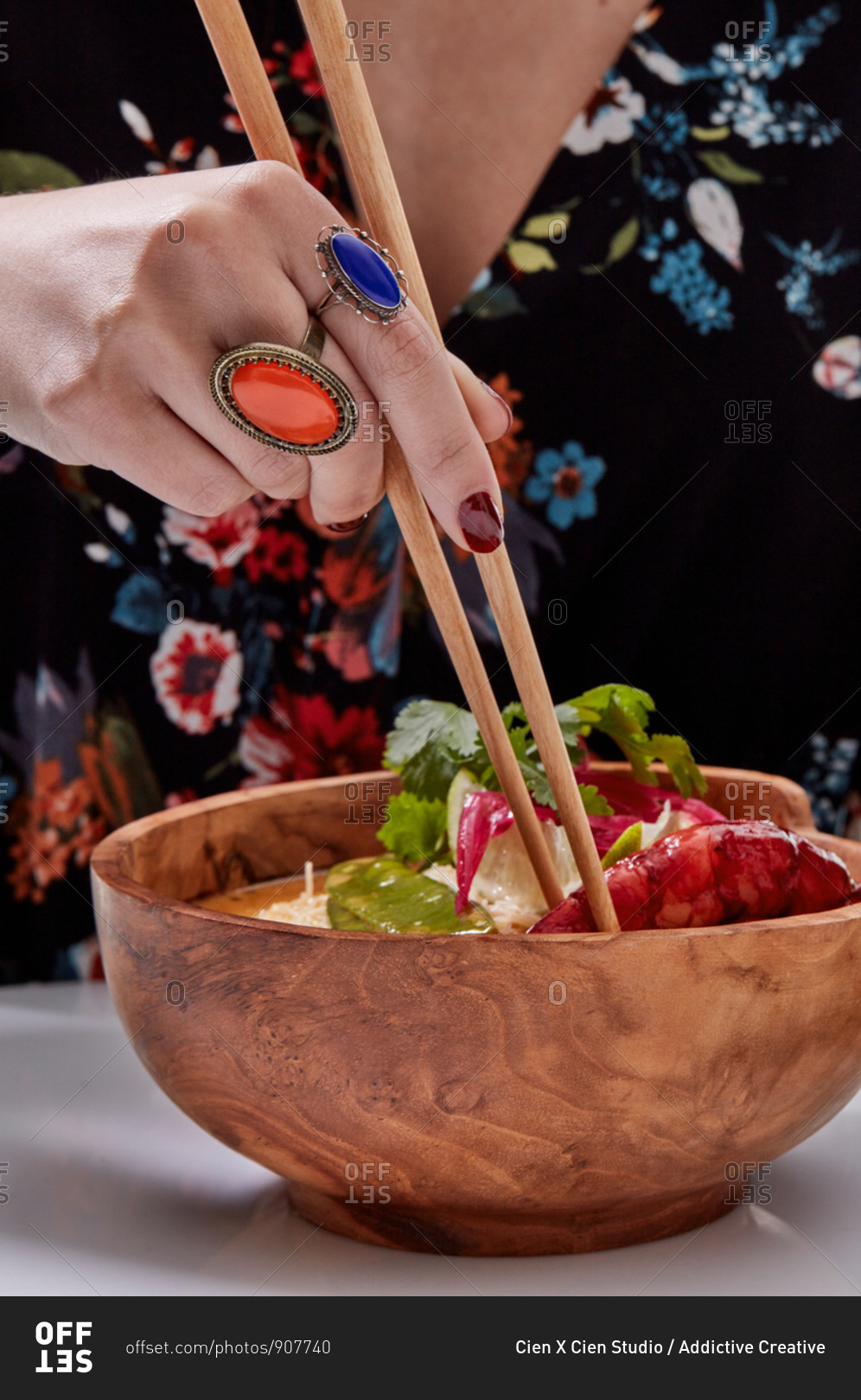 Crop hand with stylish orange and blue rings of faceless woman holding wooden chopsticks while eating Asian noodle bowl