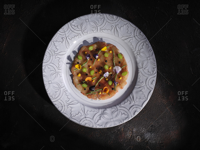 Top view of chopped condiments and petals on flat mushroom cap served on round plate on black surface