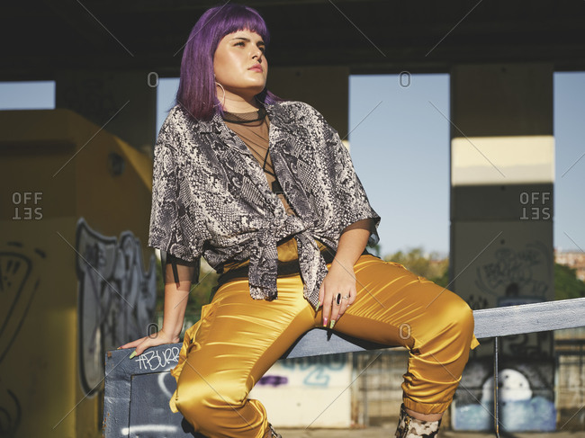 Stylish woman with bright purple hairstyle in yellow pants on metal fence in covered city station in bright day looking away