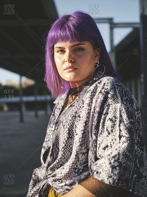 Stylish woman with bright purple hairstyle in yellow pants on metal fence in covered city station in bright day looking at camera